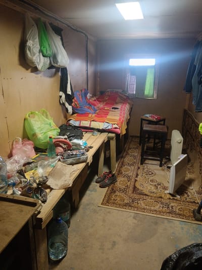 The image depicts a sparsely furnished room in Яльгелево, Russia. The room contains a bed with red and yellow blankets, a small window with curtains, and a wooden bench laden with various items, including plastic bags, bottles, and tools. A pair of hiking boots sit on the floor near the bench. A small rug covers a portion of the floor, and two electric heaters are positioned near the rug. Clothing hangs on the walls. The overall environment appears cluttered and utilitarian.