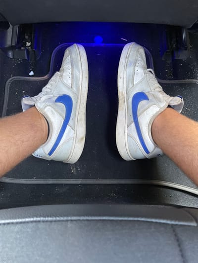 The image displays a first-person, downward-facing perspective of a person's lower legs and feet inside what appears to be the passenger footwell of a vehicle. Two male legs, visibly hairy, are extended forward with the feet resting flat on the dark carpeted floor. Each foot is encased in a white athletic shoe featuring a prominent blue Nike swoosh on the lateral side. The shoes appear worn and have some dirt accumulation, particularly on the white soles and toe boxes. Black rubber floor mats are present on the carpet, with a distinct raised edge visible horizontally across the lower third of the frame, separating the mat from the front edge of a dark car seat. The vehicle's floor is covered in visible dust and small debris. Overhead, in the upper portion of the footwell, a distinct blue light source is illuminated, casting a soft blue glow on the area directly in front of the shoes. A dark paneling, likely part of the dashboard or console, is visible on the left and right sides of the footwell. The specific city location of Mogadouro, Portugal, is not discernible from the visual information presented in the image.
