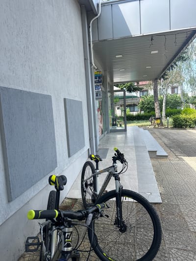 In Ovoshtnik, Bulgaria, two bicycles are parked on a paved walkway in front of a building. The building has a white, textured wall with two large, square grey panels. A covered walkway extends from the building, supported by a metal beam. Christmas lights are strung along the underside of the cover. The bicycles are parked side-by-side, with the front bicycle's handlebars and front wheel prominently in the foreground. Both bicycles have black frames with yellow accents on the seats and handlebars. Behind the building, a street with more paved surfaces and grassy areas is visible, along with trees and residential buildings.