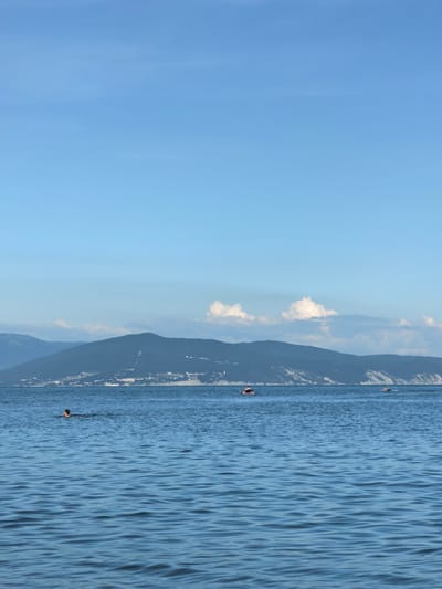 The image captures a serene coastal scene in Myskhako, Russia, dominated by a vast expanse of blue sea under a clear, pale blue sky. The horizon is marked by a range of dark, undulating mountains, partially obscured by a thin layer of clouds and mist. The slopes of these mountains are dotted with small structures, indicating a settlement.
In the foreground, the sea is a deep blue, with gentle ripples creating a textured surface. Several individuals are visible in the water. To the left, a person appears to be swimming, with only their head and shoulders above the surface. Further out, closer to the mountains, two individuals are seen on what appear to be inflatable rafts or paddleboards. One raft is orange, with a person on it, while another is smaller and further to the right, with a person on it as well. In the far distance, the faint outline of what might be boats or other vessels can be seen near the coastline. The overall atmosphere is tranquil, suggesting a warm, sunny day at the sea.