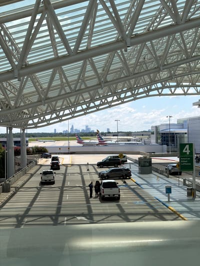 The image captures a view from within an airport terminal, looking out onto the tarmac and a street below. The foreground is dominated by the distinctive geometric structure of the airport's roof, composed of white metal beams and glass panels that cast dramatic shadows onto the scene. Below the roof, a multi-lane roadway is visible, with several vehicles. On the left side of the roadway, a white pickup truck is parked, and further down, a dark SUV. Two individuals, one in a dark uniform and another in casual clothing, stand near the white pickup truck. Further along the roadway, other vehicles are present, including a black SUV and a silver sedan.

Beyond the roadway, the airport tarmac is visible. Two airplanes are parked, one with "American" branding and another with a red and blue striped tail. In the distance, the skyline of Charlotte, North Carolina, is visible, with several tall buildings indicating the city center. The sky is a clear blue with scattered white clouds. A green sign with white lettering indicating "4 American Etihad Lufthansa" is mounted on a pole on the right side of the image. The overall impression is a sunny day at a busy transportation hub.