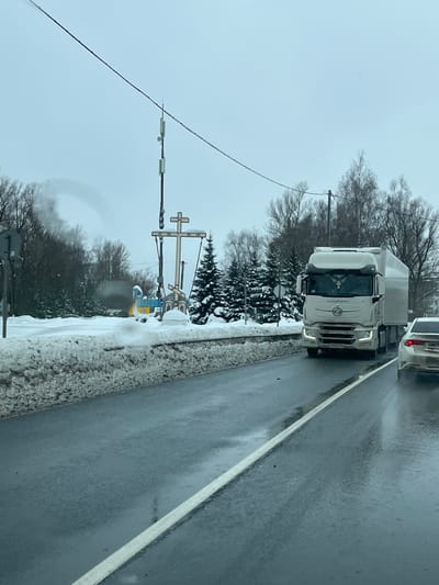 The image presents a winter roadside scene in Rogachovo, Russia, under an overcast sky. A dark, wet asphalt road occupies the foreground, delineated by a solid white line on its right side. A large, irregular bank of compacted snow runs along the left edge of the road, separating it from the adjacent terrain. In the midground, a white semi-truck, comprising a tractor unit and a trailer, is in motion, heading towards the viewer on the right side of the road with its headlights active. A portion of a lighter-colored sedan is visible directly behind the semi-truck. To the left of the road, a significant religious monument is present, featuring a large, snow-dusted wooden Orthodox cross with two smaller crosses suspended from its horizontal bar. Beneath the main cross, additional smaller religious iconography or shrines are visible. Immediately adjacent to this monument, a blue and yellow sign with Cyrillic text, confirming the location as "Рогачёво" (Rogachovo), stands. A tall utility pole, equipped with multiple communication antennas, rises behind the cross. The background consists of a dense arrangement of bare deciduous trees and snow-covered evergreen trees, extending towards the horizon under a grey, cloud-filled sky. No individuals are discernible in the scene.