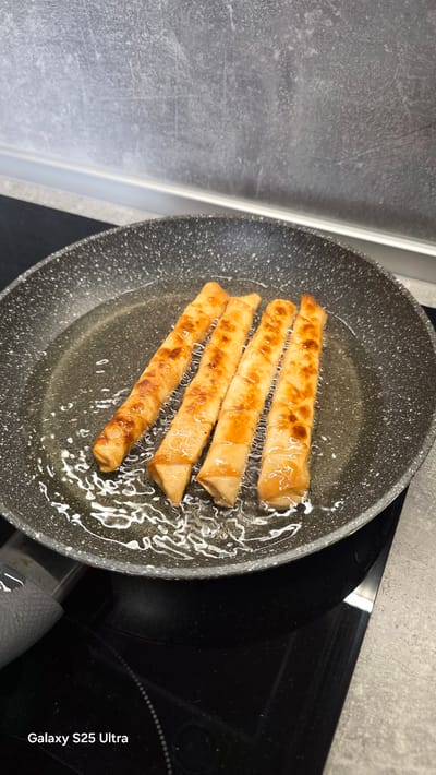Four golden-brown spring rolls are being fried in a black non-stick frying pan filled with oil. The spring rolls are arranged diagonally in the pan. The pan is on a black induction stovetop. The background consists of a grey tiled wall with a white backsplash. A grey handle of the frying pan is visible on the left side. The lower right corner of the image displays "Galaxy S25 Ultra" text. There is no indication of the city of Ezerovo, Bulgaria in the image.