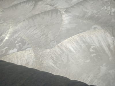 The image is a close-up, low-angle shot of a textured, metallic wall with a faint sheen, possibly a decorative finish in a building. The wall exhibits a pattern of curved, brush-stroke like indentations that create a rippling or striated effect, catching the light in varying degrees. The overall color palette is a muted silver and gray, with some areas appearing brighter due to reflections, and others darker where the texture casts subtle shadows. In the lower portion of the frame, a dark, soft object, likely upholstery or fabric, partially obscures the bottom edge of the wall, suggesting it might be the armrest or side of a piece of furniture. There are no people visible in the image. The scene is abstract and lacks any specific urban context or identifiable landmarks to place it in Petrovka, Kyrgyzstan, or any other city.