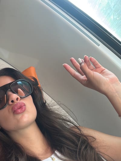 A tilted, close-up photograph captures a woman in a relaxed pose, likely within a vehicle, during daylight hours. The image features a partial view of her face and one hand, set against a light-colored interior background and a bright window.
In the lower right section, the woman's face is visible from the nose down. She is wearing large, dark sunglasses. Her lips are pursed, suggesting a "kissy face" or playful pout, and are adorned with pink lipstick. Her long, dark hair is spread around her head. A small, orange, rectangular object, possibly a cushion or headrest, is partially visible behind her head.
Her right arm and hand extend towards the upper left. Her fingers are slightly splayed, displaying fingernails with white tips, consistent with a French manicure style. A shiny, light-colored bracelet or watch is on her wrist.
The background consists of light-grey or white interior panels, possibly from a modern train, bus, or car, given the structure visible. In the upper left corner, a bright, sun-drenched window is prominent, indicating a clear, sunny day. The outside view is overexposed and blurred, emphasizing the intense natural light.
The image portrays a casual and perhaps playful moment, suggesting the woman is relaxing or posing for a photo while traveling or during leisure time. The bright sunlight confirms it is daytime. No text is visible in the image. The location context of Cagnes-sur-Mer, France, implies she may be enjoying a sunny day in that region.