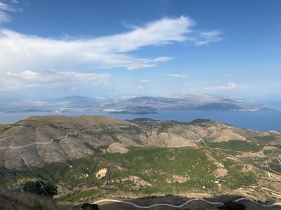 This media file depicts a panoramic, high-angle view of a mountainous, coastal landscape, likely in Greece. The scene is dominated by rugged, undulating hills and mountains covered in sparse vegetation, ranging from dry, brown grasses to patches of darker green scrub and trees. Winding, unpaved roads snake across the hillsides, suggesting a remote or rural setting.

In the distance, a body of calm, blue water, likely the sea, is visible, dotted with islands or peninsulas. The distant coastline features more rugged terrain and hints of structures or settlements. The sky above is a vibrant blue, with scattered white clouds, some appearing soft and wispy, others more dramatic.

The time of day appears to be daytime, likely late morning or early afternoon, given the bright sunlight casting shadows across the landscape. The weather seems pleasant, with a clear sky and good visibility, though some haze is present on the horizon.

There are no people or specific objects clearly discernible in the image, emphasizing the natural landscape. The overall mood is one of vastness, natural beauty, and tranquility. The terrain and the presence of the sea strongly suggest a Mediterranean coastal location, consistent with the provided location context of Kassopaia Municipal Unit, Greece. No visible text is present in the image.