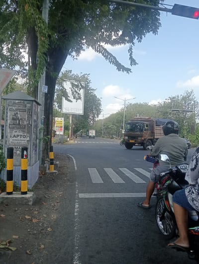 The image depicts a street scene in Banjarkemantren, Indonesia. In the foreground, two individuals are seated on a motorcycle at a pedestrian crossing. The person in the front, wearing a grey shirt and shorts, has a black helmet. The person behind them, wearing a blue shirt and jeans, is partially visible.

On the left side of the road, a utility pole stands next to a kiosk covered in posters. Yellow and black striped bollards mark the edge of the sidewalk. In the background, a brown truck with a tarp-covered bed is driving on the road. Further down the street, trees line both sides, and utility poles with streetlights are visible. A traffic light with a red signal is illuminated overhead. The sky is blue with scattered clouds. The overall impression is of a typical urban street during daylight hours.