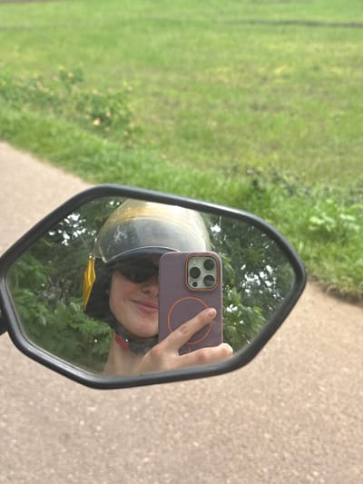 In Karandugoda, Sri Lanka, a person wearing a yellow and black helmet and sunglasses is captured in the reflection of a motorcycle's side mirror. They are holding a purple smartphone, angled to take a selfie. The background shows a blurred green grassy field and a dirt path. The image appears to have been taken outdoors on a sunny day.