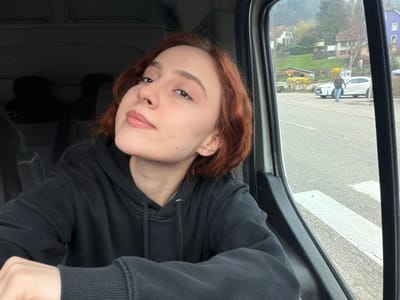 An image captured in Plaine, France, shows a young adult female seated inside a vehicle, occupying the left half of the frame. She has reddish-brown, wavy hair and fair skin, with a small silver stud earring visible in her right ear. She is wearing a black hooded sweatshirt and looks towards the camera with a neutral expression, her lips slightly parted. The vehicle's interior appears dark, with dark-colored seats visible in the background.
Through the vehicle's window, the external environment is visible. An asphalt road occupies the mid-ground, featuring white pedestrian crossing stripes in the lower right. A light-colored SUV is parked on the roadside. A male individual, dressed in dark clothing and jeans, walks away from the camera on the road, positioned near the parked SUV. The background features a green hillside with multiple residential buildings, including one with a purple section, and scattered trees with light green foliage and yellow flowering shrubs under an overcast sky.