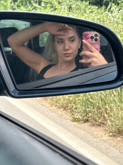 In this medium shot, a young woman is seen reflected in a car's side mirror. She has blonde hair, fair skin, and is wearing a black tank top. Her right hand is raised, holding a pink phone with a decorative case, and her left hand is resting on her forehead. The reflection shows her face looking slightly downwards, with a neutral expression.
The setting appears to be outdoors, likely during the daytime, as indicated by the bright natural light. The background visible in the mirror is a blur of green foliage and a hint of a road or path. This suggests the car might be parked or moving through a rural or semi-rural area. The phone's case is pink with a floral pattern and has a prominent camera bump. The woman's fingernails are painted white. The overall mood is casual and personal, possibly a moment captured while traveling or taking a break. There is no visible text in the image. The location context is Parvomai, Bulgaria.