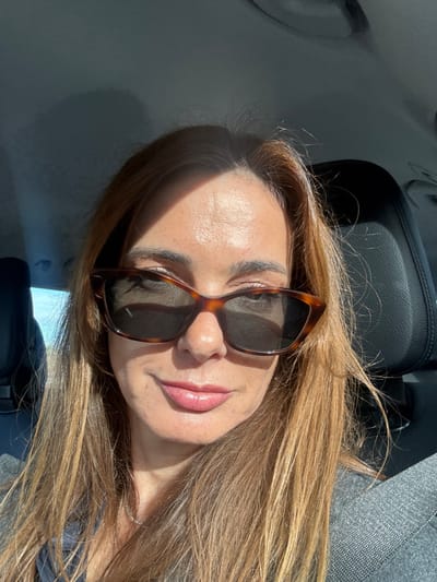 The image displays a close-up selfie of an adult female taken inside a vehicle, purportedly located in Alcobaça, Portugal. The subject has light brown hair with auburn tones, extending past her shoulders. She wears brown tortoise-shell cat-eye sunglasses with dark lenses. Her lips are closed in a slight smile and appear to have a light pink, glossy finish. A delicate, silver-toned necklace is visible around her neck. She is dressed in a dark blue or navy top, covered by a grey, textured garment, possibly a blazer or jacket. Intense direct sunlight illuminates the left side of her face and hair, creating distinct shadows. In the background, to the left of the subject, a bright blue sky or distant outdoor area is visible through a window. To her right, a black, leather-like vehicle headrest is present. The vehicle's interior ceiling appears dark. The subject is looking directly at the camera.