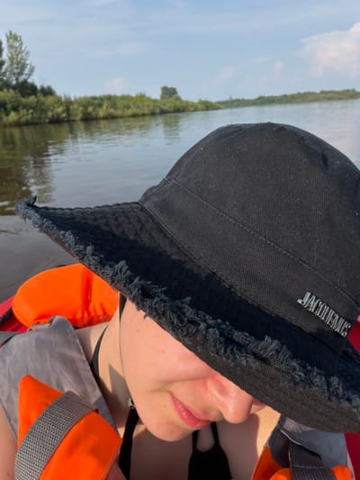 A close-up shot shows a person wearing a black bucket hat and an orange life vest on a body of water. The hat has a frayed edge and a silver "JACQUEMUS" logo on the side. The person's face is partially visible, with their head tilted down. The background shows a calm river with green foliage on the banks and a blue sky with some clouds. The scene appears to be in the city of Kalino, Russia, likely during daylight hours.