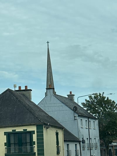 The image is a vertical, outdoor, medium shot of rooftops and a church spire under a cloudy sky. The scene appears to be in a town or city, with the architecture suggesting an urban environment. In the foreground, there's a dark grey roof with multiple chimneys. Behind it, a lighter grey, pointed spire with a cross at its apex extends upwards. The spire is attached to a building with a light grey sloped roof. Further in the background, a multi-story building with numerous windows, some with security bars, is visible. A large green tree with dark foliage is also present, and a lamppost is attached to the side of a building. The sky is overcast and light blue, indicating daytime. There is no visible text in the image. The overall impression is one of a slightly dreary, possibly historic urban landscape. The image's orientation is rotated 90 degrees counter-clockwise.