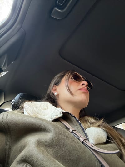 A woman in sunglasses looks upward from inside a car with a sunroof. She wears an olive green jacket with white fur trim. Her lips are pursed slightly. A beige purse strap is visible across the jacket. The car's interior is predominantly black, with the sunroof taking up a significant portion of the frame. The background is blurred, suggesting movement.