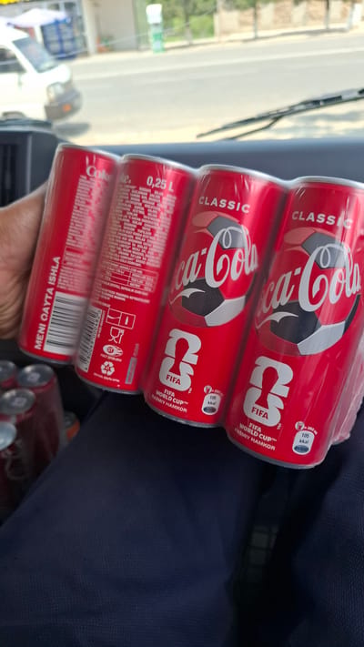 This media file captures a close-up, angled shot of a six-pack of Coca-Cola cans, prominently displaying a "FIFA World Cup" theme. The cans are predominantly red with white lettering, featuring the iconic Coca-Cola logo along with the FIFA World Cup branding. A soccer ball design is also integrated into the Coca-Cola logo on each can.

The scene appears to be inside a vehicle, suggested by the presence of a windshield wiper and a partial view of the car's interior and the exterior environment through the window. The lighting suggests it's daytime, possibly with bright sunlight contributing to the overall illumination. The background outside the window shows a blurred outdoor scene with greenery, hinting at a location with natural elements.

One hand is visible in the lower part of the frame, holding the pack of cans. The cans themselves are the primary subject, with detailed text visible on the labels, including nutritional information and product details in what appears to be Uzbek language. Text like "MENI QAYTA ISHLA" (which translates to "Recycle me") and "Coca-Cola" are clearly legible, alongside the "FIFA World Cup" and "RASMIY HAMKOR" (Official Partner) designations.

The overall impression is a casual, everyday moment of someone holding a purchase, likely for personal consumption or transport. There are no strong emotional cues, and the focus is on the product itself and its branding.
