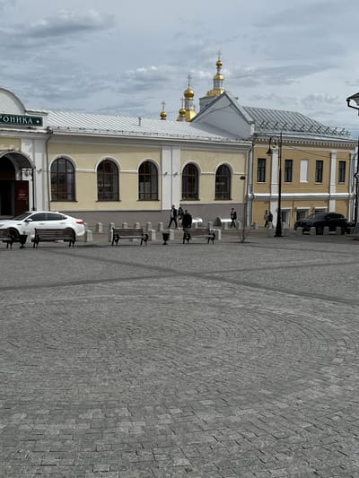 The image shows a street scene in Arzamas, Russia. The street is paved with gray cobblestones in a curved pattern. On the left side of the image is a multi-story building with a beige facade, arched windows, and a gray metal roof. Golden domes and crosses are visible on top of the building, indicating a religious structure. A sign with Cyrillic writing is visible on the building's entrance. Along the building's facade, there are benches and several people walking or sitting. A white car is parked near the building. On the right side, another building with a similar architectural style is partially visible. The sky is overcast with gray clouds.