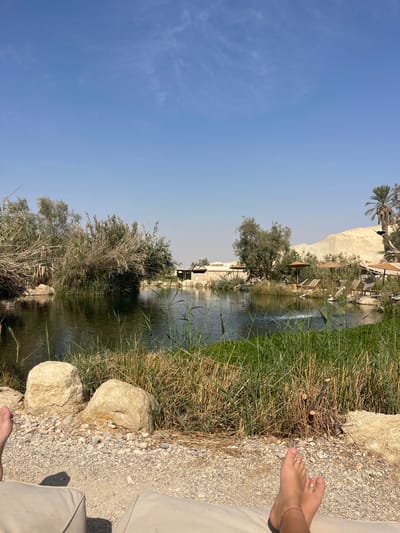 The image captures an outdoor recreational area, likely a desert oasis or resort, within the Arava Tichona Regional Council, Israel, under a clear blue sky. In the immediate foreground, two human feet are visible, resting on a light-colored cushioned surface. The right foot features two toe rings (on the second and third toes) and a thin ankle bracelet. The ground beneath the feet consists of light-colored gravel and scattered larger stones.
Beyond the immediate foreground, a tranquil body of water, resembling a pond or small lake, occupies the midground. Its surface reflects the blue sky, and dense green and brown reeds and vegetation line its banks, particularly on the left. A small water aerator or fountain is visible on the right side of the water, creating minor ripples.
In the background, across the water, several low-rise buildings with light-colored walls and dark roofs are present. Adjacent to these structures, multiple vacant lounge chairs are arranged beneath brown-canopied umbrellas. Further in the distance, a prominent light-colored desert mountain or hill range rises on the right side of the frame, with a single palm tree visible nearby. The scene is brightly illuminated by direct sunlight, indicating a clear day.