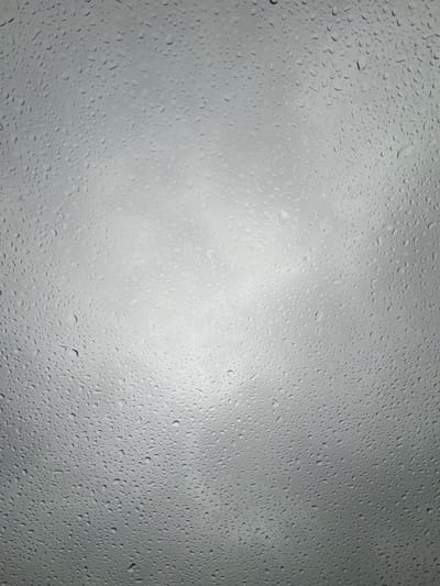 This media file is a close-up, abstract photograph of water droplets on a surface, likely glass, given the context of rain. The droplets vary in size and distribution, creating a textured pattern across the frame. The background appears to be a bright, overcast sky, indicated by the diffuse light and subtle variations in shade. There are no people, objects, or distinct scene elements visible, making the image purely textural and atmospheric.
The photograph captures the visual effect of rain on a window or similar transparent surface. The lighting suggests it is daytime, possibly mid-day, due to the bright but diffused light, common on a cloudy or rainy day. The weather is clearly rainy. While the specific location is Jõhvi, Estonia, this is not visually evident from the image itself. The image evokes a sense of calmness, introspection, or perhaps melancholy, often associated with rainy weather. There is no visible text in the image.