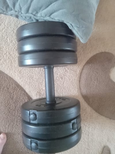 A black dumbbell with adjustable weights rests on a textured beige carpet. A light blue blanket is draped over the top of the dumbbell. The dumbbell consists of a central handle with a textured grip, surrounded by stacked weight plates. The weight plates are secured with black clips. A person's thumb is visible in the lower left corner of the image, near the bottom weight plates. The scene appears to be indoors, likely in a domestic setting. No specific location in the city of Terebovlia, Ukraine, can be determined from the image.