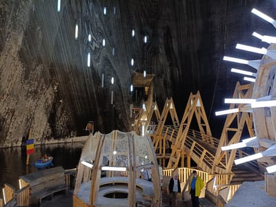 The image depicts the interior of the Salina Turda salt mine in Turda, Romania. The cavernous space is illuminated by numerous hanging cylindrical lights and light strips integrated into wooden structures. In the foreground, a wooden, multi-tiered structure with netting and seating is visible. To the left, a body of dark water is present, with a small blue boat carrying at least two individuals and a Romanian flag on a pole. Further into the background, complex wooden walkways and elevated platforms, also adorned with lighting, ascend the cavern walls. Several people are observed walking on these platforms and near the foreground structures. The salt mine walls, characterized by their stratified, textured appearance in varying shades of gray and brown, form the backdrop of the scene.