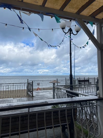 The image shows an overcast view of the Baltic Sea from an outdoor cafe or restaurant seating area in Pionersky, Russia. The foreground is occupied by a wet wooden deck with railings, suggesting recent rain. In the middle ground, a sandy beach is visible, separated from the seating area by a metal fence. A life preserver ring is hanging on the fence. Further back, the sea stretches to the horizon under a cloudy sky. A street lamp with decorative flags and string lights is positioned in the middle ground, adding to the ambiance of the seaside establishment. Benches are scattered throughout the seating area.