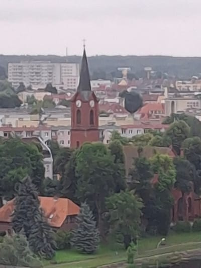 The image shows a hazy, aerial view of a town, likely Pila, Poland, focusing on a church with a tall, dark steeple topped with a cross. The church is constructed of red brick and features two clock faces on its tower. Surrounding the church are numerous trees, some with dense foliage and others appearing as evergreens. In the background, there are various buildings, including apartment blocks and residential houses with red tiled roofs. The overall atmosphere is overcast, suggesting a cloudy day. There are no visible people or discernible actions taking place within the scene.