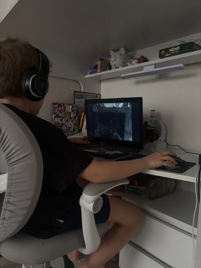 An individual with light brown hair, wearing black over-ear headphones with a lightning bolt logo, a black and grey short-sleeved shirt, and black shorts, is seated at a white desk. The person is positioned with their back to the camera, engaged in playing a video game on an open black laptop. The laptop screen displays a dark, possibly industrial or ruined environment from a first-person perspective, featuring a circular reticle. The individual's left hand rests on the laptop keyboard, while the right hand operates a black computer mouse on a dark mousepad. To the right of the laptop, a white and red instrument resembling a small microscope is visible. Above the desk, a white floating shelf holds a white ceramic piggy bank with black spots, a small red lidded container, a green box labeled "MEMO," and other small items. A white LED light bar is mounted beneath this shelf. The person is seated on a grey ergonomic office chair with light grey armrests. The walls are white. This scene is located in Krasnaya Zarya, Russia.