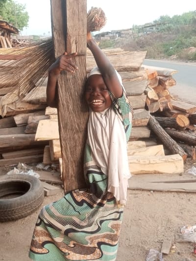 A young girl smiles while holding onto a wooden post. Her arms are raised, one clutching a bundle of sticks. She wears a patterned dress and a light-colored headscarf. Behind her is a stack of wooden planks and logs. A tire rests on the ground near the post. The background shows a road and some distant buildings, suggesting an outdoor setting perhaps near a lumberyard. The lighting appears soft and natural.