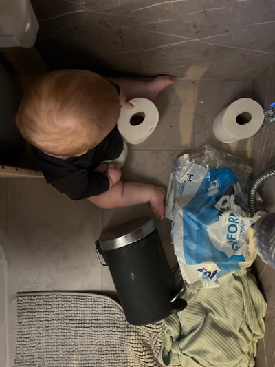 A baby sits on a tile floor in a bathroom near a roll of toilet paper. A second roll sits on the floor near the shower head. A blue plastic bag of toilet paper rests next to a black metal garbage can, which is next to a light-colored towel and a grey bathmat. The baby wears a black shirt and a diaper.