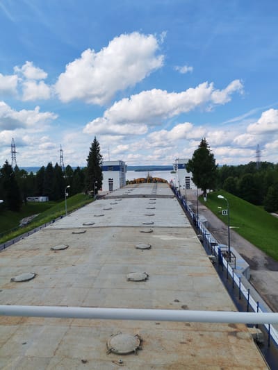 An overhead perspective shot showcases a vast, weathered, and metallic surface dotted with circular, rusty manhole covers. This expansive surface appears to be the roof of a large industrial structure, possibly a barge or a dam. A white railing runs horizontally across the foreground, framing the view of the metallic expanse. To the right of the metallic surface, a steep, grassy embankment slopes downwards, bordered by a blue and white railing. A paved walkway runs alongside the embankment, featuring a lamppost with a sign displaying the number "9".
Further in the background, two modern white buildings with blue accents stand prominently. Between them, a tall, dark green coniferous tree reaches towards the sky. Beyond the buildings and the tree-lined horizon, a wide body of water is visible, reflecting the bright blue sky dotted with fluffy white clouds. High voltage power line towers are scattered throughout the landscape, suggesting an industrial or power generation facility in the vicinity. The overall impression is one of a functional, industrial area integrated with natural elements. The location is identified as Белоусово, Russia.