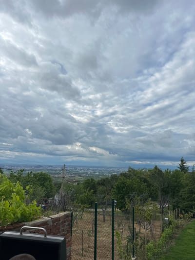The image presents a view from an elevated position overlooking what appears to be the city of Brestnik, Bulgaria. The sky is dominated by a pattern of long, streaky clouds covering most of the frame. A landscape with trees, bushes, and some sort of fenced area is visible on the right side, descending slightly into the urban area. A brick structure and vegetation can be observed in the foreground.