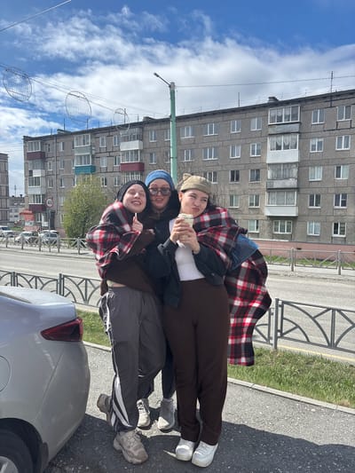 Three people are posing in an urban environment. The background consists of a gray, multi-story apartment building with multiple windows and possibly balconies. The sky is visible with patches of blue and white clouds. Power lines are present, decorated with circular decorative elements. A streetlight is also visible. The group is positioned near a curb and part of a car is visible at the bottom of the frame. The setting is identified as Kachkanar, Russia.