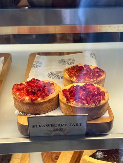 The image shows three strawberry tarts displayed in a glass case at a bakery in Santa Fe, Costa Rica. The bakery is located at the coordinates 9.642, -85.165. The tarts are arranged on a wooden board lined with parchment paper and labeled "Strawberry Tart 2.500" in a handwritten sign. The tarts are topped with a generous amount of sliced strawberries and a creamy filling. The case is made of glass and has a metal frame. The tarts are arranged in a row, with the leftmost tart facing the viewer, the middle tart slightly angled, and the rightmost tart facing away from the viewer. The scene is lit by artificial light.