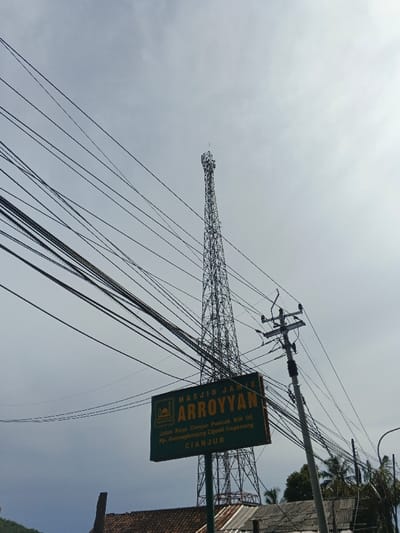 The image features a communication tower against a pale sky, cluttered with numerous overhead power lines. A green sign labeled "MASJID JAINE ARROYYAN" stands in the foreground, providing directions to Jalan Raya Cianjur Puncak KM 06, Kp. Gununglanjung Cijedil Cugenang, Cianjur. A utility pole with transformers is positioned to the right of the sign. The base of the tower and the sign are partly obscured by rooftops of nearby buildings.