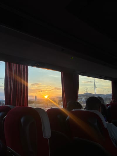 The image captures the interior of a passenger bus, viewed from a seat towards the front-right, during either sunrise or sunset. The bus's dark ceiling is visible at the top, featuring recessed lighting or vents and a small green indicator light on the left. Red and black passenger seats, some with white headrest covers, occupy the foreground and midground. Deep red, pleated curtains are partially drawn across the windows on the left side of the bus.
Outside the left window, the sun is intensely bright, appearing as a prominent disc with a starburst effect, positioned above a black traffic light pole. A red "STOP" sign is partially visible below the traffic light. Further in the background, a light-colored building with partially legible Greek text (including "ΕΛΛΑΔΑ") is visible, against a sky transitioning from orange-yellow at the horizon to lighter blue, with scattered clouds and distant hills.
Outside the right window, the sky displays similar hues with scattered clouds. A large power line pylon (trestle tower) is visible on a distant hill, along with multiple streetlights on poles. Buildings and urban structures are present in the middle distance. A white vehicle is partially visible on the far right side of the road.
Inside the bus, strong rays of sunlight penetrate the cabin, creating lens flare and illuminating parts of the interior and passengers. Several passengers are visible from behind, seated. One individual in the mid-right has dark hair and wears a white collared shirt, appearing to be observing a small electronic device on their lap. The scene is located within the Municipal Unit of Lamia, Greece.