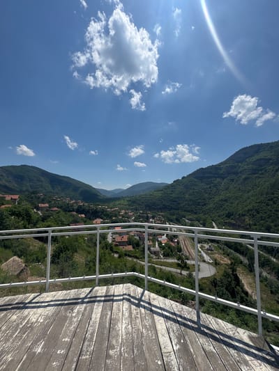 The image captures a panoramic view of a mountainous valley on a clear, sunny day, from what appears to be an elevated observation point in the city of Zasele, Bulgaria. In the foreground, a weathered wooden deck with extensive peeling grey and white paint is visible, extending from the bottom of the frame to approximately one-third up. Distinct shadows of a white metal railing are cast across the wooden planks, suggesting the sun is high and to the left. The railing itself, constructed from white metal, forms the immediate barrier, featuring a top handrail, a lower support bar, and several slender vertical posts.
Beyond the railing, a deeply verdant valley unfolds, flanked by steep, densely forested mountains on both the left and right sides. The slopes are covered predominantly with various shades of green trees and shrubs. Nestled within the valley and along the lower slopes, a village is visible, characterized by numerous houses with distinct orange and red-tiled roofs, interspersed among the trees. Winding roads, some paved and some appearing unpaved, snake through the village and along the contours of the landscape.
To the right side of the valley, a railway line with multiple tracks is clearly discernible, curving through the terrain, adjacent to a paved road. A small, light-colored structure with a dome, possibly a chapel or monument, is situated near the railway tracks. The mountains recede into the distance, exhibiting a lighter, hazy green hue. The sky above is a bright, clear blue, populated by several scattered white cumulus clouds of varying sizes. A prominent lens flare, appearing as a rainbow-like arc, is visible in the upper right quadrant of the sky, indicating the direct sunlight. No individuals or specific activities are present in the scene.