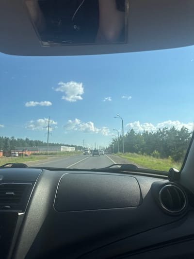 The image depicts a view from inside a car driving on a highway. The dashboard is in the foreground, with a black textured surface and air vents visible. Beyond the windshield, a paved road stretches ahead, with other vehicles in motion. On either side of the road, there are grassy areas and dense trees. Power lines and streetlights are also present. The sky is blue with scattered white clouds. There is no discernible indication of the location being the city of Vasil'evka, Russia.