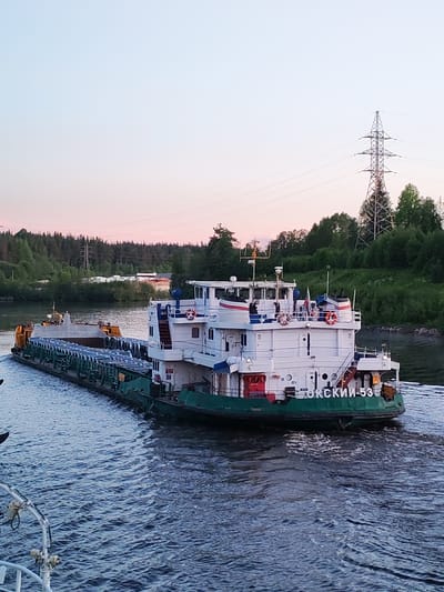 A large cargo vessel navigates on a wide river or canal during twilight hours. The sky above is a blend of soft pastel blues and pinkish-orange, suggesting either sunrise or sunset.
The vessel consists of a prominent white superstructure with multiple decks, a control bridge, and red detailing, sitting on a green hull. Russian flags are visible on the superstructure, along with red and white lifebuoys. The Cyrillic text "ОКСКИЙ-53" (OKSKIY-53) is clearly visible on the stern of the white section, identifying the ship. Attached to the vessel is a long, flat cargo barge laden with numerous uniformly sized cylindrical objects, possibly large pipes or industrial coils, arranged lengthwise. The vessel generates a visible wake as it moves through the water.
The banks of the waterway are densely covered with green trees and foliage. In the distance, on the far bank, a tall metal electricity pylon stands against the sky, with power lines stretching across. The image is taken from another vessel, with a white railing partially visible in the bottom left corner. The scene depicts tranquil transit on a waterway, likely in Russia, consistent with the provided context of "Depo, Russia."