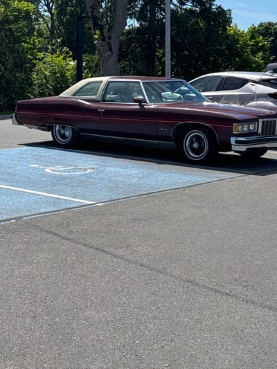 In a parking lot in Tabasalu alevik, Estonia, a maroon 1970s sedan is parked in a disabled parking spot marked with a blue surface and a white wheelchair symbol. The car, which has a white vinyl roof, is facing right. A silver SUV is parked to the right of the sedan. The background consists of trees and a lamppost. The ground is paved with asphalt.