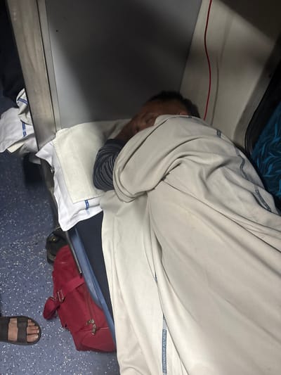 A person is asleep on a train. They are covered in a light gray cloth, with their head resting on a white pillow. A red bag is near their legs. A sandal-clad foot is visible. The train interior has a blue patterned seat and gray walls.