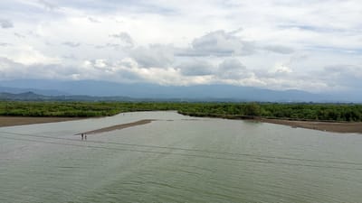 The image shows a wide, murky green river flowing through a lush, tropical landscape near Baroh Lancok, Indonesia. The riverbanks are lined with dense green vegetation, likely mangrove forests, which stretch towards a backdrop of hazy, distant mountains. The sky is overcast with dramatic, grey clouds. In the foreground, two figures are visible wading in the shallow water near a sandy bank. The river appears to be a significant waterway, possibly connecting to the sea, given the proximity to the coast and the type of vegetation. Two power lines stretch horizontally across the lower portion of the image, indicating human infrastructure in the vicinity.