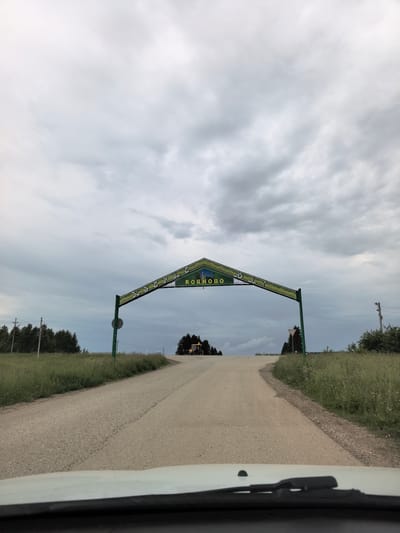 The image shows an overhead archway sign with the Russian text "КОЯНОВО" in yellow letters against a green background. The arch spans a paved road that curves to the right. The sky is overcast with gray clouds. On the left side of the road, there is a field of tall green grass and a line of trees. On the right side, there is also grass and trees, and further in the distance, a yellow bulldozer is visible. The photograph appears to be taken from the perspective of someone inside a vehicle, looking forward at the sign.