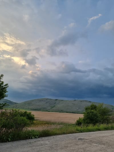 The image depicts a landscape in Aldomirovtsi, Bulgaria, under a cloudy sky with some golden light breaking through. A mountain ridge runs along the right side of the frame, with visible text on its slope. At the base of the mountain, there is a field of dry vegetation. A dirt road runs along the right edge of the image, bordering green vegetation. The sky occupies a significant portion of the image, filled with various cloud formations.
