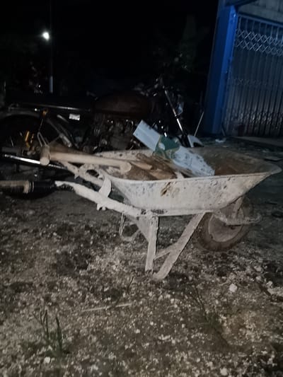 The image depicts a nighttime scene in Sendangrejo, Indonesia, showing a motorcycle and a wheelbarrow. The motorcycle appears to be on its side, with its front wheel facing the left and its exhaust pipe visible. The wheelbarrow is positioned in front of the motorcycle and contains various items, including what appear to be wooden planks and a white object. The ground is covered in gravel and dirt. A blue metal gate is visible in the background to the right, and a street light can be seen illuminating the scene from the left. The overall lighting is dim, suggesting it is dark outside.