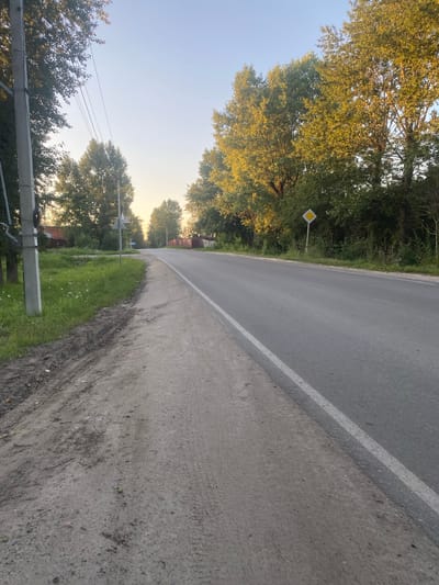The image shows a paved road with a white line marking its edge, running through a rural landscape in Semyonovskoye, Russia. The road curves gently to the left, bordered by a concrete shoulder and a grassy verge. Tall trees with lush green foliage line the left side of the road, with some trees exhibiting golden autumn colors, suggesting it's late summer or early autumn. In the distance, a yellow diamond-shaped road sign is visible amongst the trees.
Power lines stretch across the frame, supported by concrete poles, indicating the presence of a village or settlement nearby. The sky is clear and bright, with a soft, warm light indicating either early morning or late afternoon. The sun appears to be low on the horizon, casting long shadows and illuminating the leaves of the trees with a golden hue. The overall scene evokes a sense of tranquility and the quiet beauty of a rural setting. There are no people visible in the image, and the activity is that of a static landscape.