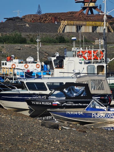 The image depicts a collection of boats docked in a rocky harbor, likely in Dudinka, Russia. Several vessels are visible, ranging from larger white ships with red life preservers to smaller, sleeker boats. One boat in the foreground has "PK 0914 RUS24" written on its hull, along with a Russian flag. Another boat bears the text "North Silver Pro 565 M" and a "MERCURY PRO XS" outboard motor. Further back, a blue and white boat displays "РКЯ04-84" and what appears to be "РЫБООХР" in Cyrillic script.

In the background, a large industrial crane towers over stacks of logs, suggesting a logging or timber processing facility. A power line pylon is also visible on a hill, hinting at the infrastructure of the area. The scene is illuminated by bright sunlight, indicating it is daytime. The sky is clear and blue, suggesting fair weather. The rocky shoreline and the presence of multiple vessels point to a working port or marina. No people are clearly visible in the frame. The overall impression is one of a functional industrial harbor.
