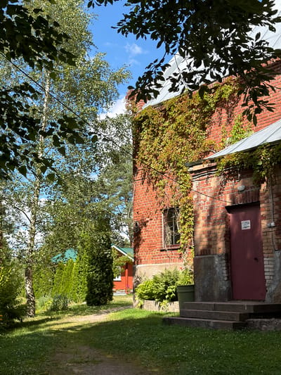 The image depicts a sunlit scene in Sevastyanovo, Russia, showcasing a brick building covered in ivy. A dark red door is visible on the right side of the building, with a sign that reads "СТОП" (STOP in Russian). Adjacent to the building are stone steps leading up to the door. To the left, a path winds through a grassy area with scattered white flowers. Lush green trees and manicured bushes line the path and surround a red house with a green roof in the background. The sky is blue with scattered white clouds. The overall impression is one of a tranquil, sun-drenched rural setting.