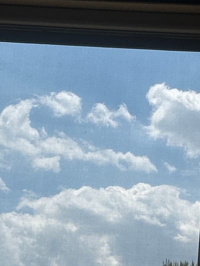 The image shows a bright blue sky with fluffy white clouds, likely captured from a window. There is a dark, horizontal bar at the top, suggesting a frame or the edge of a structure. The clouds are voluminous and appear to be cumulus clouds, scattered across the sky. Some smaller, darker objects that could be birds or insects are visible against the sky. The bottom of the frame shows a sliver of green foliage, hinting at trees or other vegetation. The scene is characteristic of an outdoor environment on a clear day. The location is specified as Militello in Val di Catania, Italy.