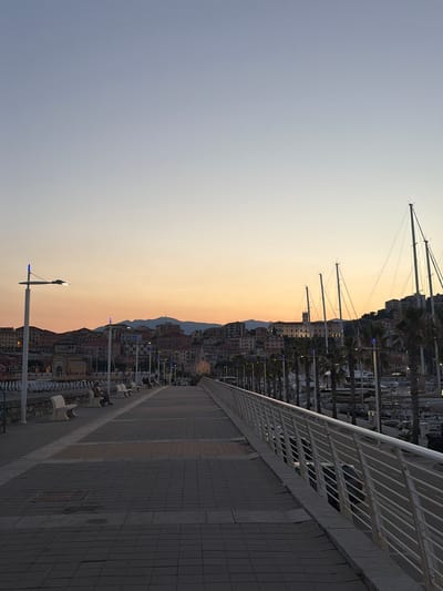 The image depicts a harbor scene at dusk in Imperia, Italy. The setting is a promenade with a tiled walkway and a white railing, leading the eye towards a cityscape nestled against a hillside. Tall sailboat masts dominate the upper left portion of the frame, silhouetted against a gradient sky transitioning from pale blue to a warm, peachy orange.
The cityscape is characterized by densely packed buildings with terracotta roofs, suggesting an Italian coastal town. Palm trees line the waterfront, and various boats are moored in the harbor, visible behind the promenade. People are seated on benches along the walkway, enjoying the evening atmosphere. Several streetlights with blue accents are visible, indicating the onset of night. The overall mood is tranquil and serene, with the warm colors of the sunset creating a peaceful ambiance. The time of day is clearly dusk, evidenced by the soft, diffused light and the transition of colors in the sky. The presence of sailboats and the architectural style of the buildings strongly suggest a Mediterranean coastal location, specifically identified as Imperia, Italy. There is no visible text in the image.
