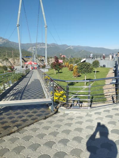 The image shows a suspension bridge in the city of Dardar, Uzbekistan, on a clear day. The bridge is made of metal with tall grey pylons supporting suspension cables. A paved walkway with a distinctive scale-like pattern leads across the bridge, lined with metal railings and planters filled with yellow and pink flowers. Beyond the bridge, a green lawn is dotted with small trees, and in the background, there are mountains. Several people are visible on a viewing platform further along the walkway, dressed in casual clothing. A red and white umbrella and tables are set up near the beginning of the bridge, suggesting a seating or refreshment area. The shadow of a person holding a phone is cast on the paved ground in the foreground, indicating the photo was taken by someone present at the location.